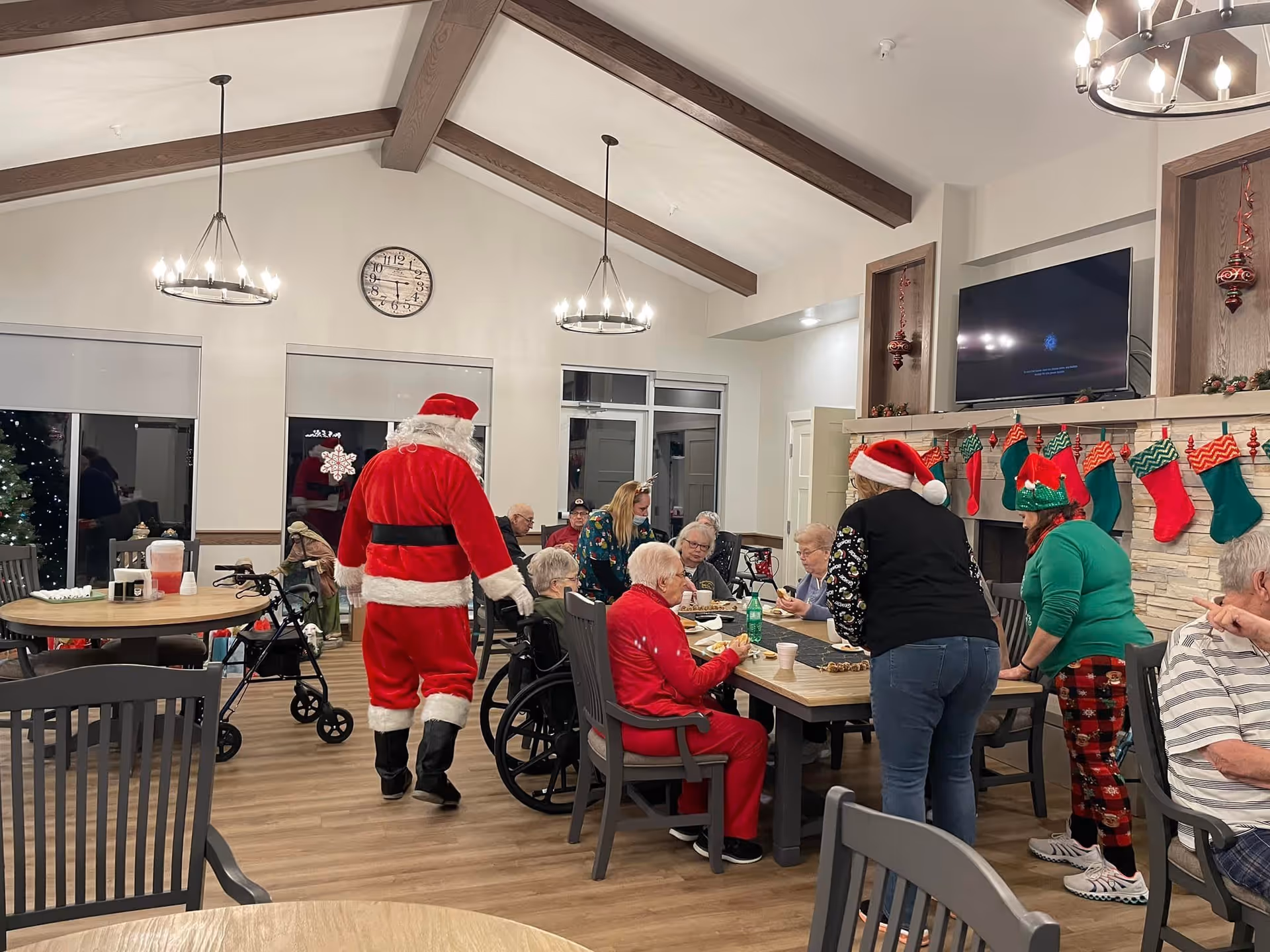 A group of elderly people and caregivers gathered around a table in a decorated common room. One person is dressed as Santa Claus, and others wear festive holiday clothing including Santa hats and elf hats. The room has wooden beams on the ceiling, a clock on the wall, a TV above a fireplace with Christmas stockings hanging, and holiday decorations.