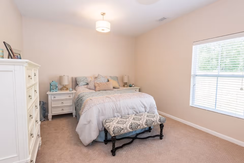 A bright bedroom with a large bed covered in light-colored bedding and multiple pillows. There are two white nightstands on either side of the bed, each with a lamp and decorative items. A patterned bench is placed at the foot of the bed. A white dresser with framed photos and decor is on the left side of the room. A large window with blinds allows natural light to fill the room.
