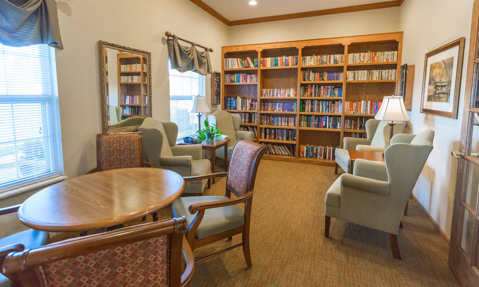 A cozy reading room with wooden bookshelves filled with books, four green upholstered armchairs, two wooden side tables with lamps, a wooden round table with four chairs, a large mirror on the wall, and two windows with blinds and valances.
