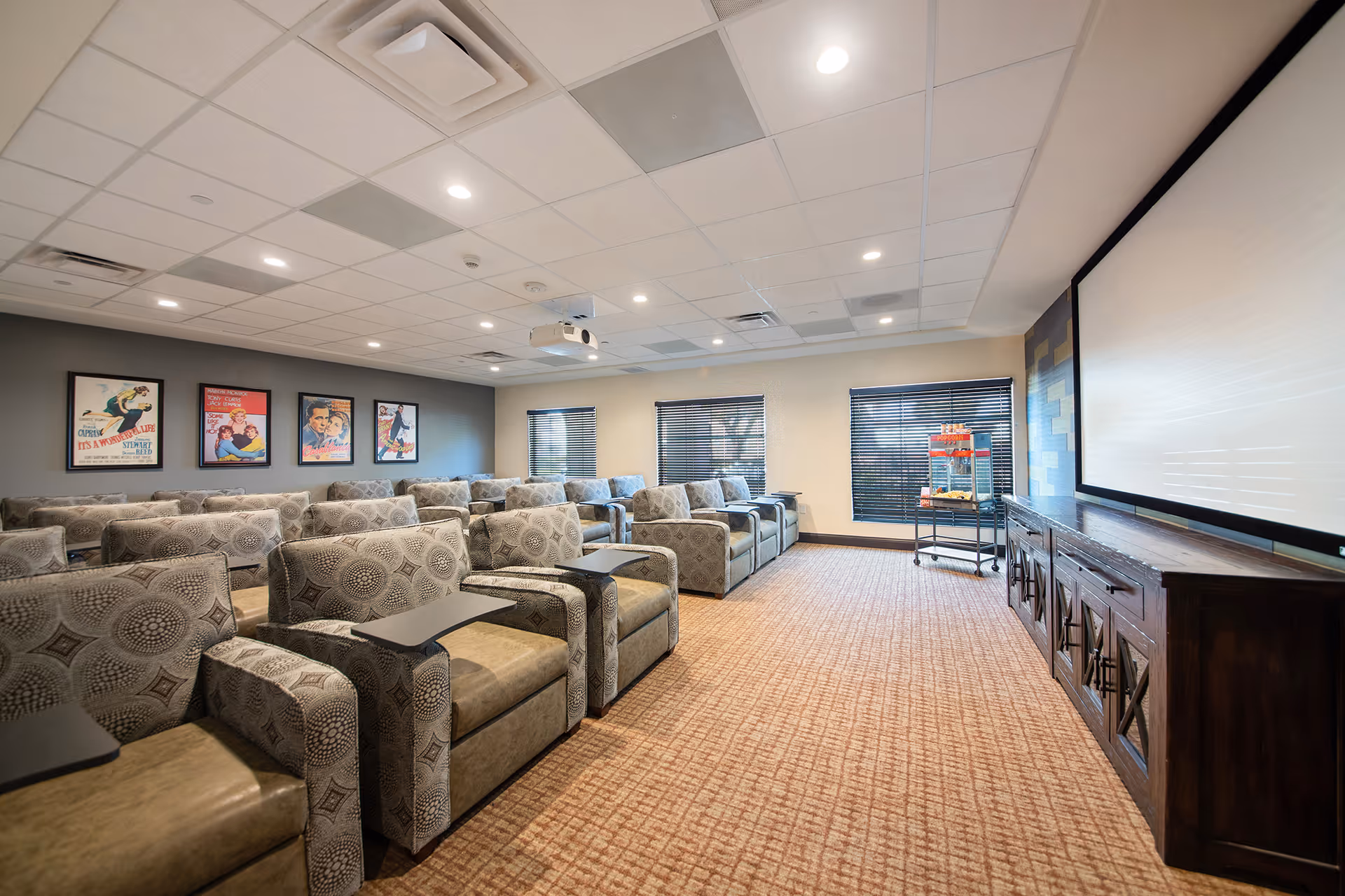 A cozy movie theater room with multiple rows of patterned armchairs equipped with small attached tables. The room has a large projector screen at the front, a popcorn machine on a cart near the windows with blinds, and several vintage movie posters on the wall. The ceiling has recessed lighting and a projector mounted in the center.