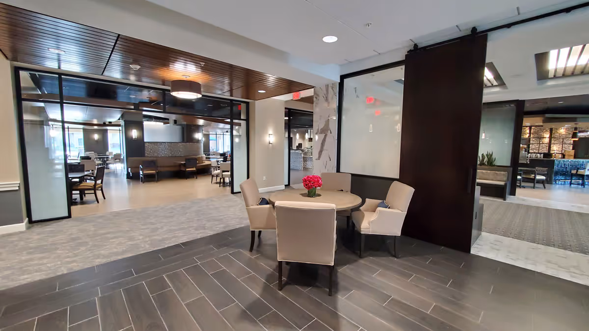 A modern senior living facility interior with a small round table and four beige upholstered chairs arranged around it. The table has a small vase with pink flowers. The space features dark wood ceiling panels, large glass partitions, and a mix of dark tile and carpet flooring. In the background, there are more seating areas and dining tables visible through the glass partitions.