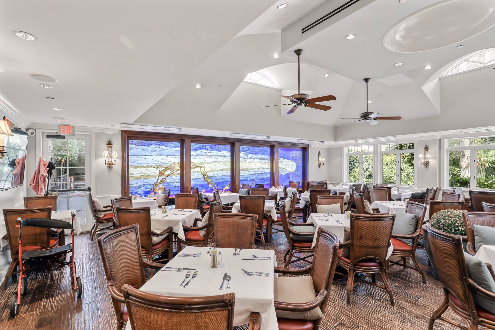 A bright dining room with multiple tables covered in white tablecloths, each set with silverware and salt and pepper shakers. The room features wooden chairs with cushions, large windows letting in natural light, ceiling fans, and a large stained glass window with blue and beige tones on one wall. A walker is visible near the entrance door.