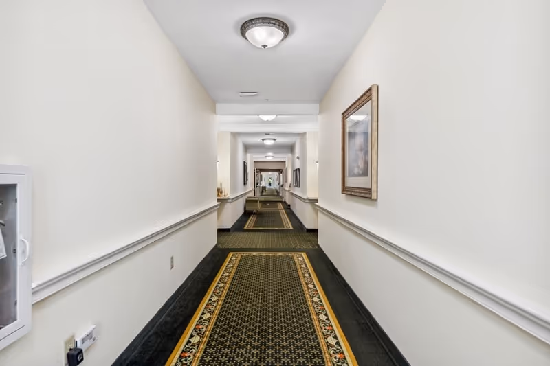 Long indoor hallway with patterned carpet runner, cream-colored walls, framed artwork on the right wall, and ceiling light fixtures. The hallway has a handrail on both sides and a fire extinguisher cabinet on the left wall.