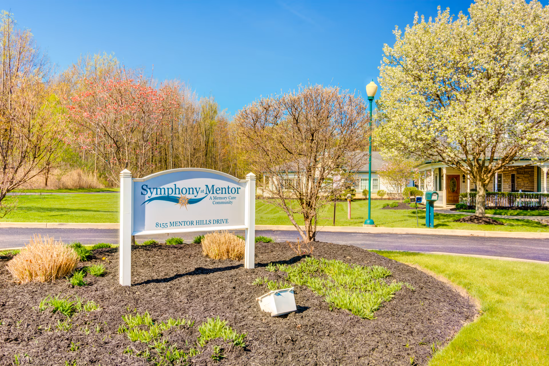 Outdoor view of Symphony at Mentor, a memory care community, showing a landscaped area with a white sign displaying the facility name and address, surrounded by trees and a building in the background under a clear blue sky.