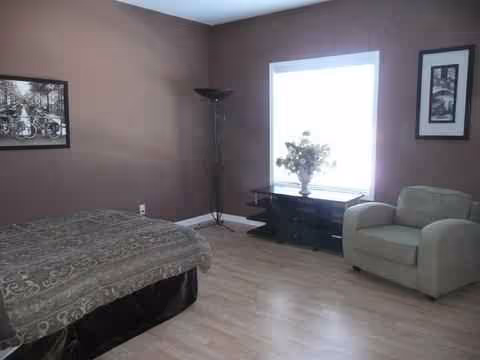 A bedroom in a senior living facility with a bed, chair, window, and side table.