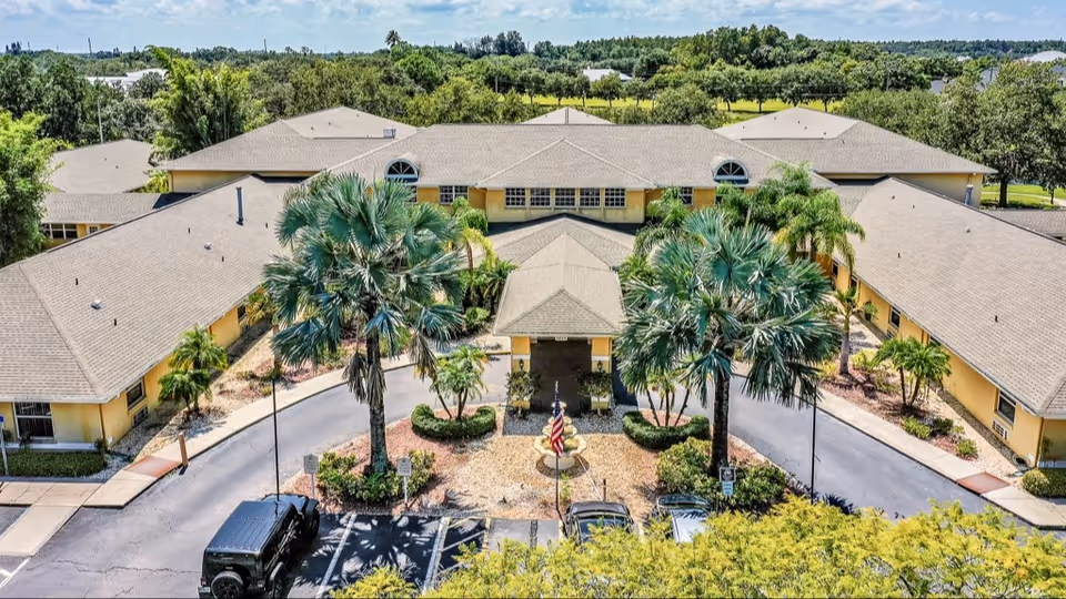 Aerial view of a single-story senior living facility with a U-shaped layout surrounded by greenery and palm trees. The building has a light yellow exterior and a gray shingled roof. There is a circular driveway with an American flag and landscaped plants in the center, and several parked cars are visible in front of the entrance.