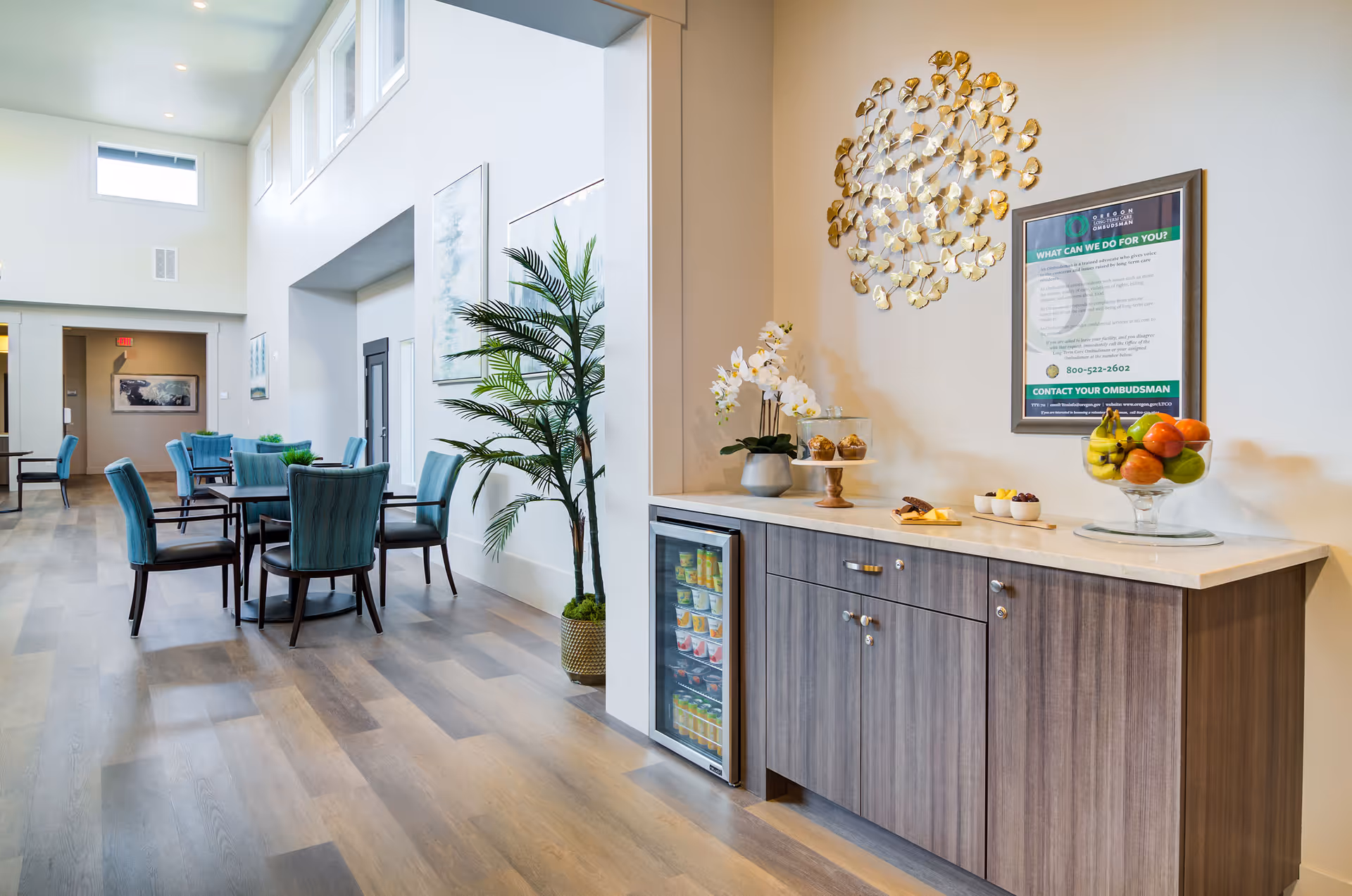 A bright and spacious common area in a senior living facility with high ceilings and large windows. There are several tables with blue upholstered chairs arranged for seating. A wooden cabinet with a small refrigerator is topped with decorative items including a bowl of fruit, a vase with white flowers, and a tiered tray with muffins. A framed informational poster is mounted on the wall above the cabinet. A tall potted plant is placed near the tables, and the floor is covered with wood-style flooring.