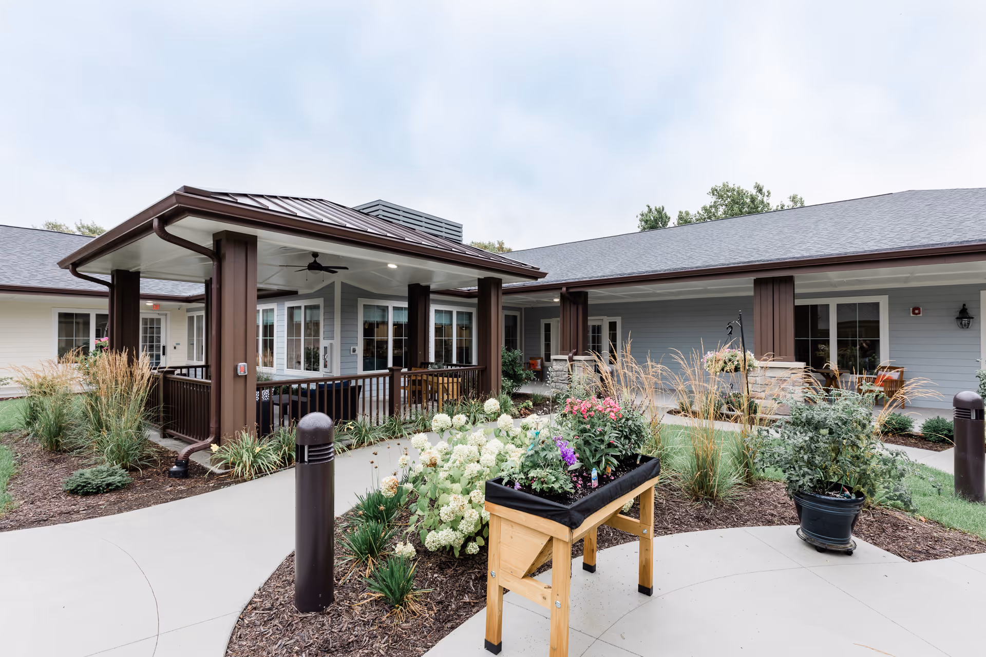 Landscaped courtyard with flowers and a raised planter in front of a single-story memory care building's covered porch.