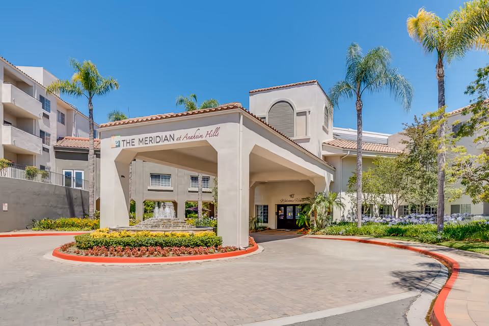 Exterior view of The Meridian at Anaheim Hills senior living facility entrance with a covered driveway, palm trees, landscaped flower beds, and a water fountain under a clear blue sky.