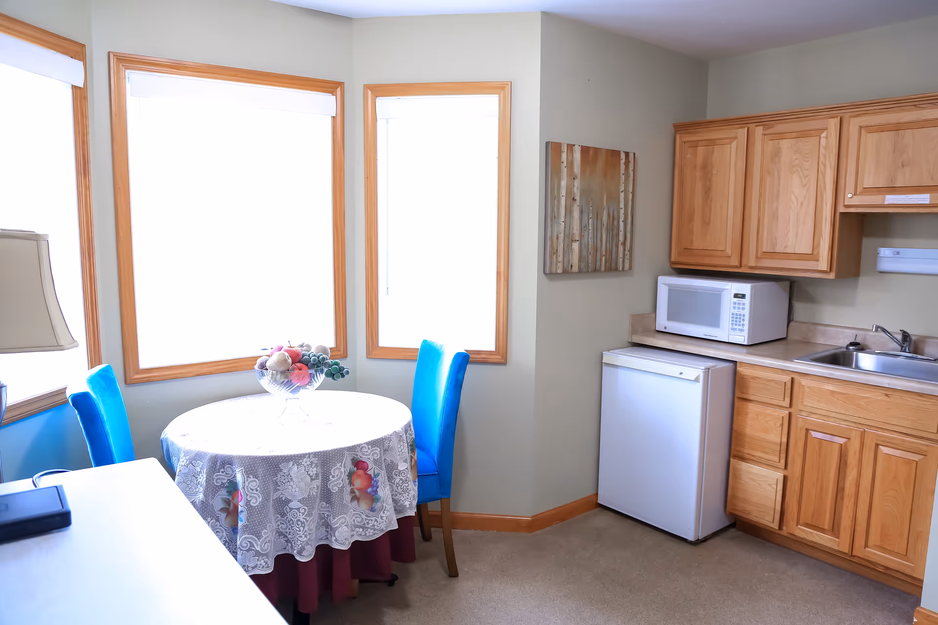 Small kitchenette and dining nook with a round table and blue chairs next to wooden cabinets, a mini fridge, microwave and sink.