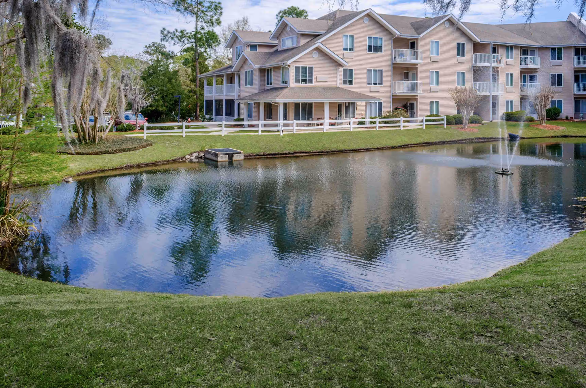 View of a multi-story residential building with beige siding and white balconies, situated behind a pond with a water fountain. The area is surrounded by green grass, trees, and a white fence along the edge of the pond under a partly cloudy sky.