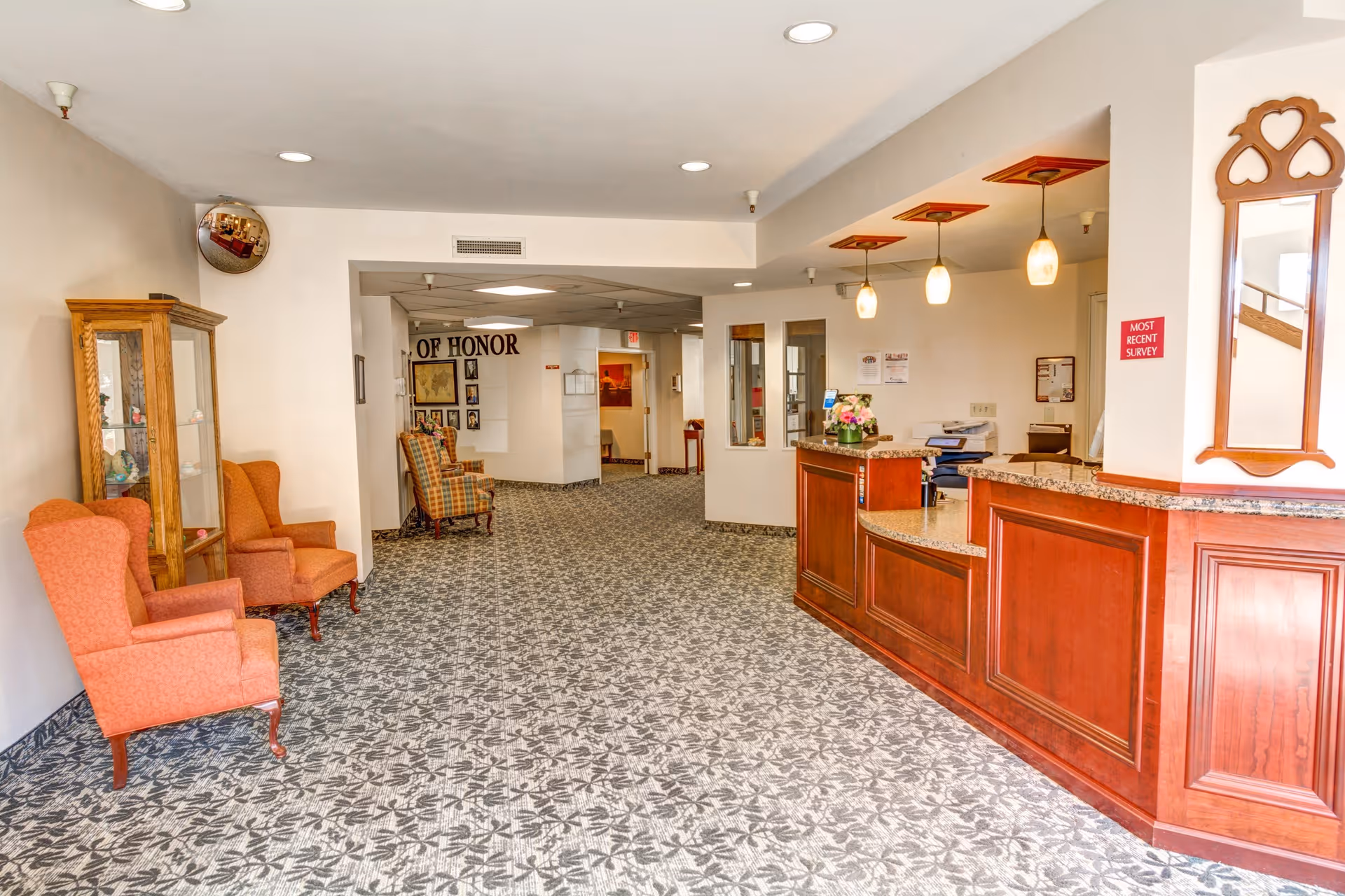 Interior view of a senior living facility reception area with a wooden front desk on the right, three hanging pendant lights above it, and a mirror on the wall. On the left side, there are two orange upholstered armchairs and a glass display cabinet. The floor is covered with patterned carpet, and in the background, there is a hallway with additional chairs and a wall display labeled 'OF HONOR'.