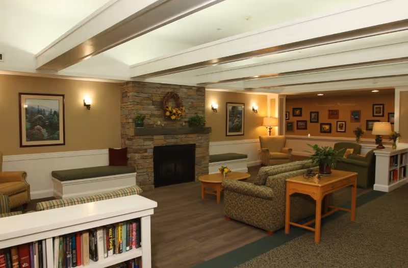A cozy living room area in a senior living facility featuring a stone fireplace decorated with a wreath and plants. The room has multiple seating options including armchairs, sofas, and built-in benches with cushions. There are framed pictures on the walls, a wooden coffee table with a small flower arrangement, and a bookshelf filled with books. The lighting is warm with wall sconces and table lamps.
