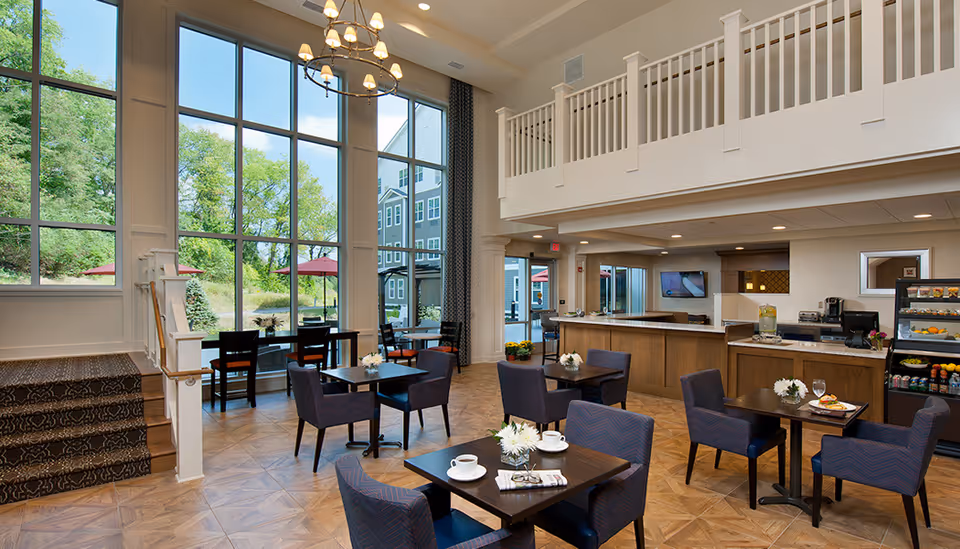 Bright two-story dining/lounge area with tables and chairs, large floor-to-ceiling windows overlooking greenery, and a service counter.