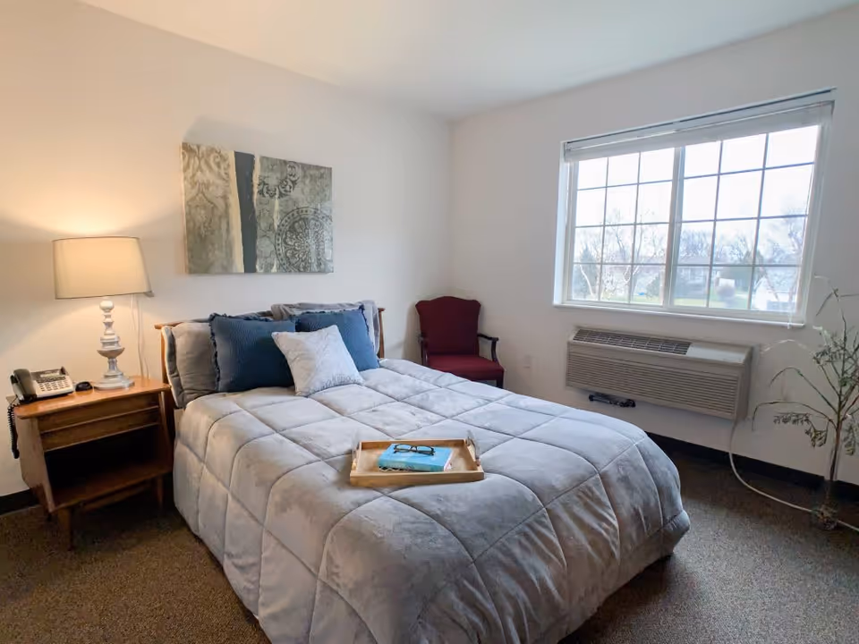 A cozy bedroom with a neatly made bed featuring gray bedding and blue and white pillows. A wooden nightstand with a lamp and a telephone is beside the bed. A red armchair is placed near a large window with white blinds, and a wall-mounted air conditioning unit is below the window. A decorative painting hangs on the wall above the bed, and a small plant is in the corner near the window.