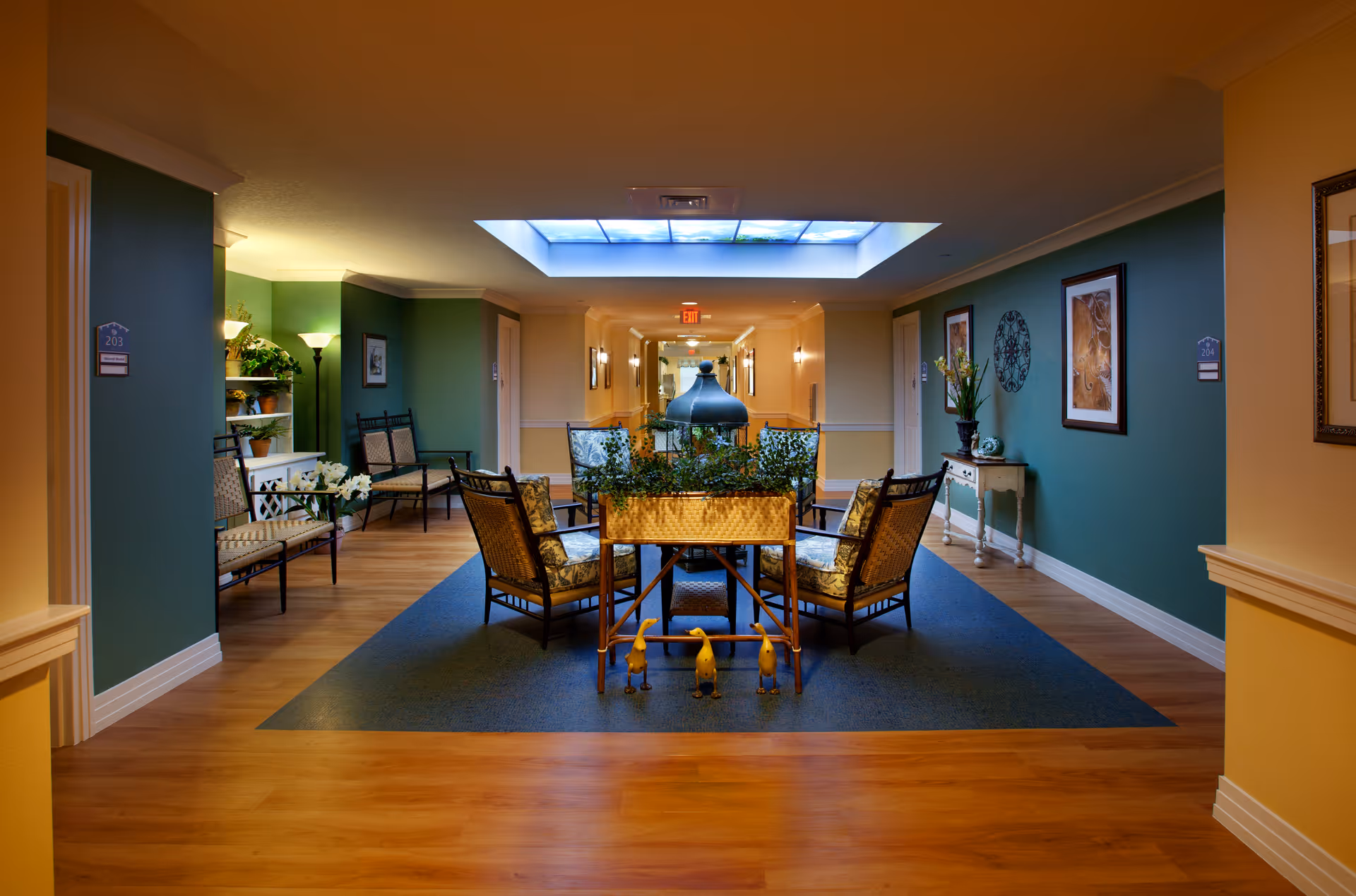 A cozy seating area in a senior living facility hallway with several cushioned chairs arranged around a central planter with greenery. The walls are painted green and beige, decorated with framed artwork and a decorative wall piece. The floor is wood with a blue rug under the seating area. A skylight with a sky design is on the ceiling, and there are room number signs on the walls.