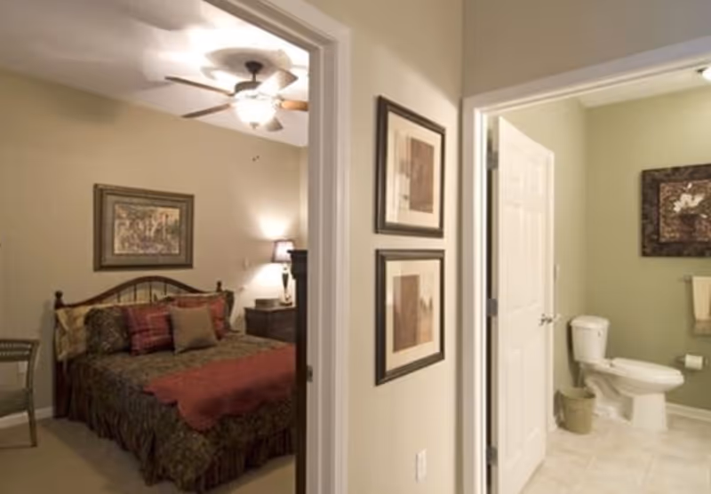 View of a bedroom with a bed covered in patterned bedding and multiple pillows, a nightstand with a lamp, and framed artwork on the wall. Adjacent to the bedroom is a bathroom with a toilet, towel rack, and framed artwork on the wall.