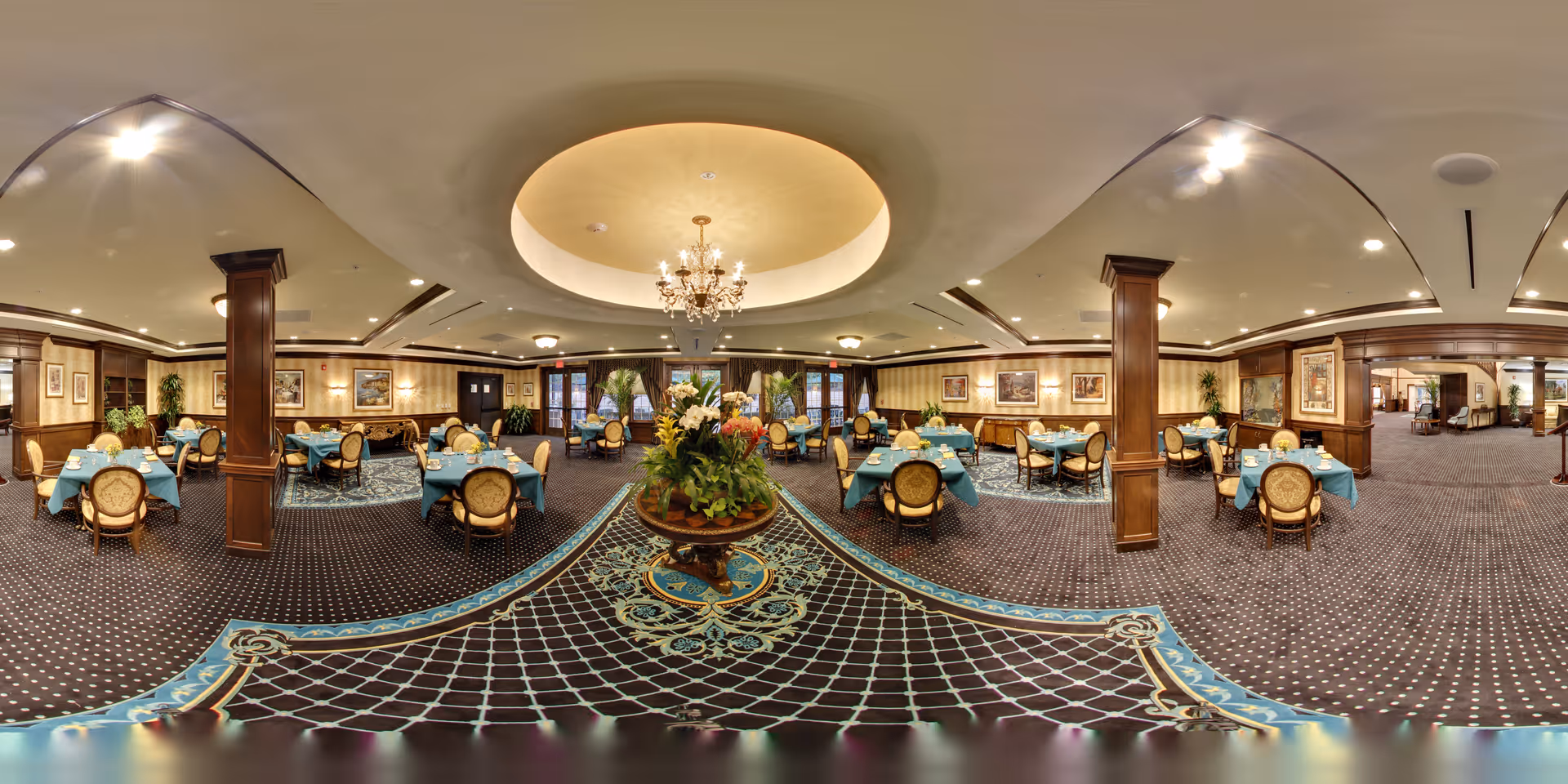 A spacious, elegant dining room with multiple round tables covered in blue tablecloths and set with white napkins and glassware. The room features dark wood paneling, framed artwork on the walls, patterned carpet, and a central chandelier hanging from a recessed ceiling. There is a large floral arrangement on a round table in the center of the room.