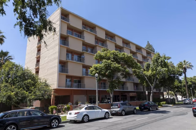 Exterior view of a multi-story residential building with balconies, surrounded by trees and parked cars along the street under a clear blue sky.