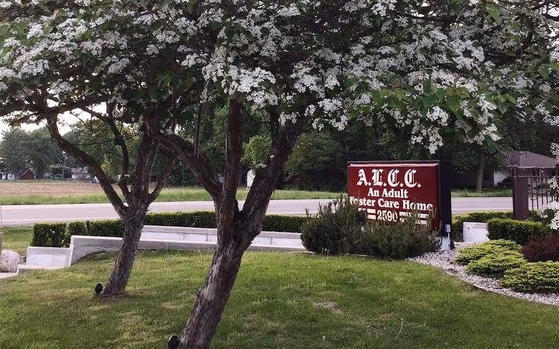 Blooming trees and a landscaped lawn by a roadside red sign reading "A.L.C.C. An Adult Foster Care Home".