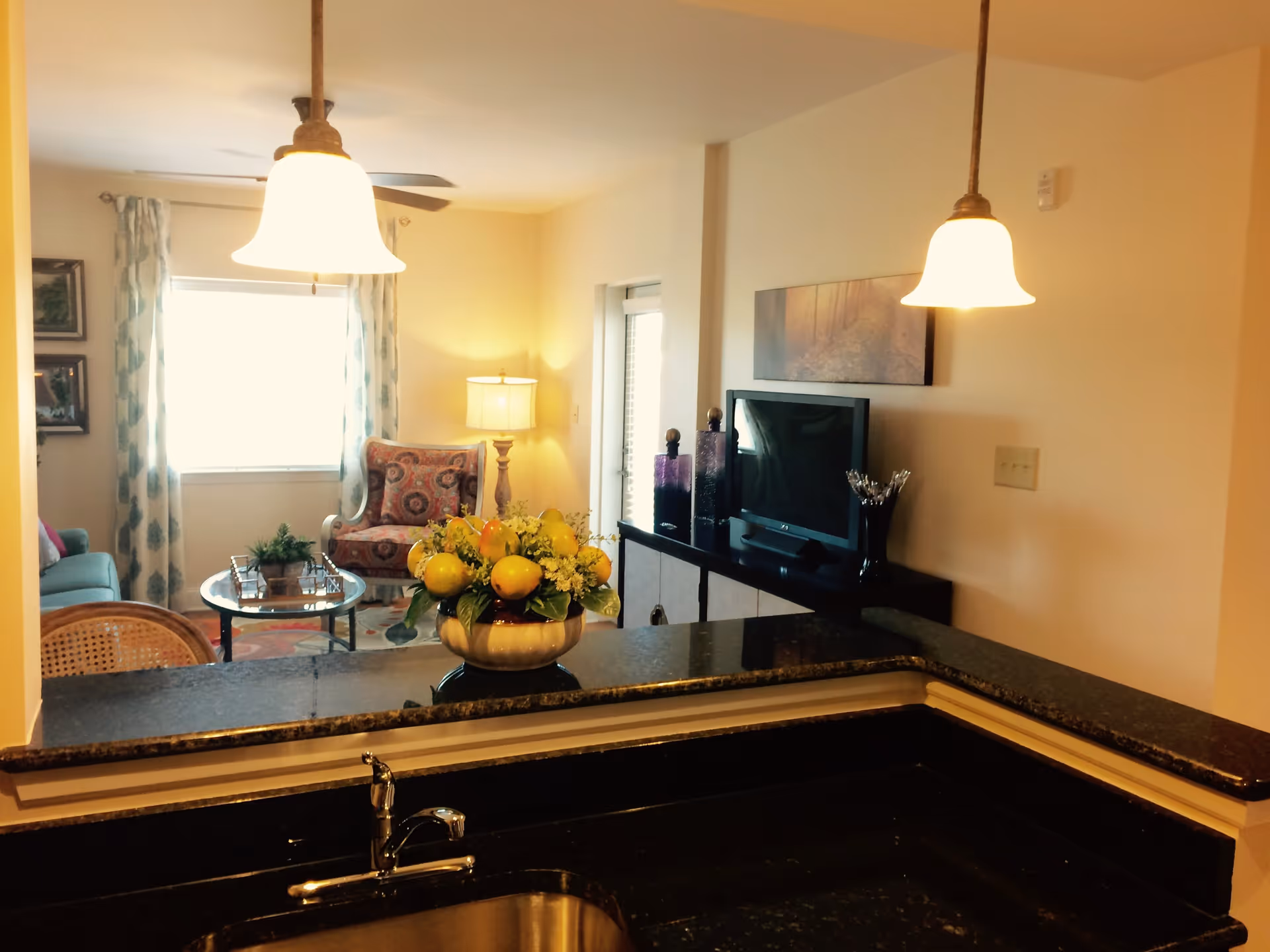 View from a kitchen sink area looking into a living room with a patterned armchair, a sofa, a glass coffee table, a TV on a stand, and a lamp. Two pendant lights hang over the kitchen counter, and a decorative bowl with yellow fruit and flowers sits on the counter.
