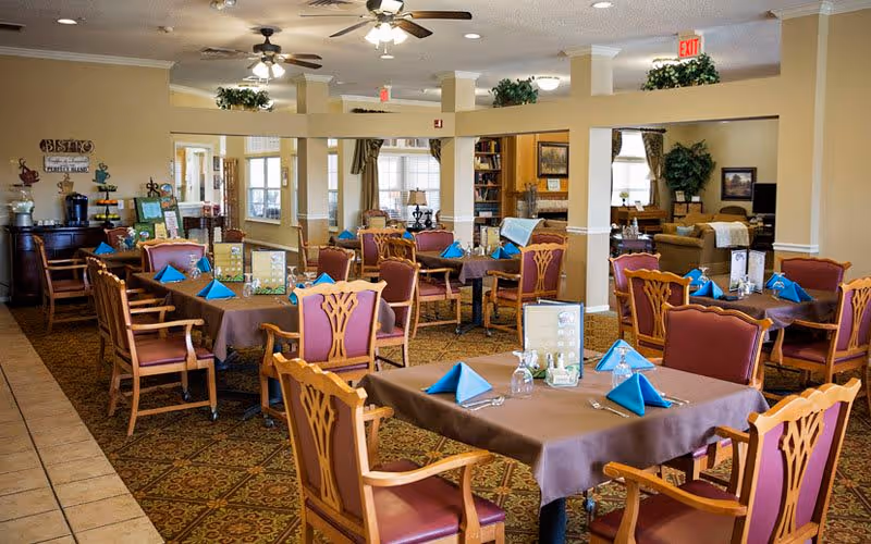 A dining room in a senior living facility with several tables covered with brown tablecloths and set with blue folded napkins, glasses, and silverware. The room has wooden chairs with red cushions, ceiling fans with lights, and beige walls. In the background, there is a cozy living area with sofas and plants.