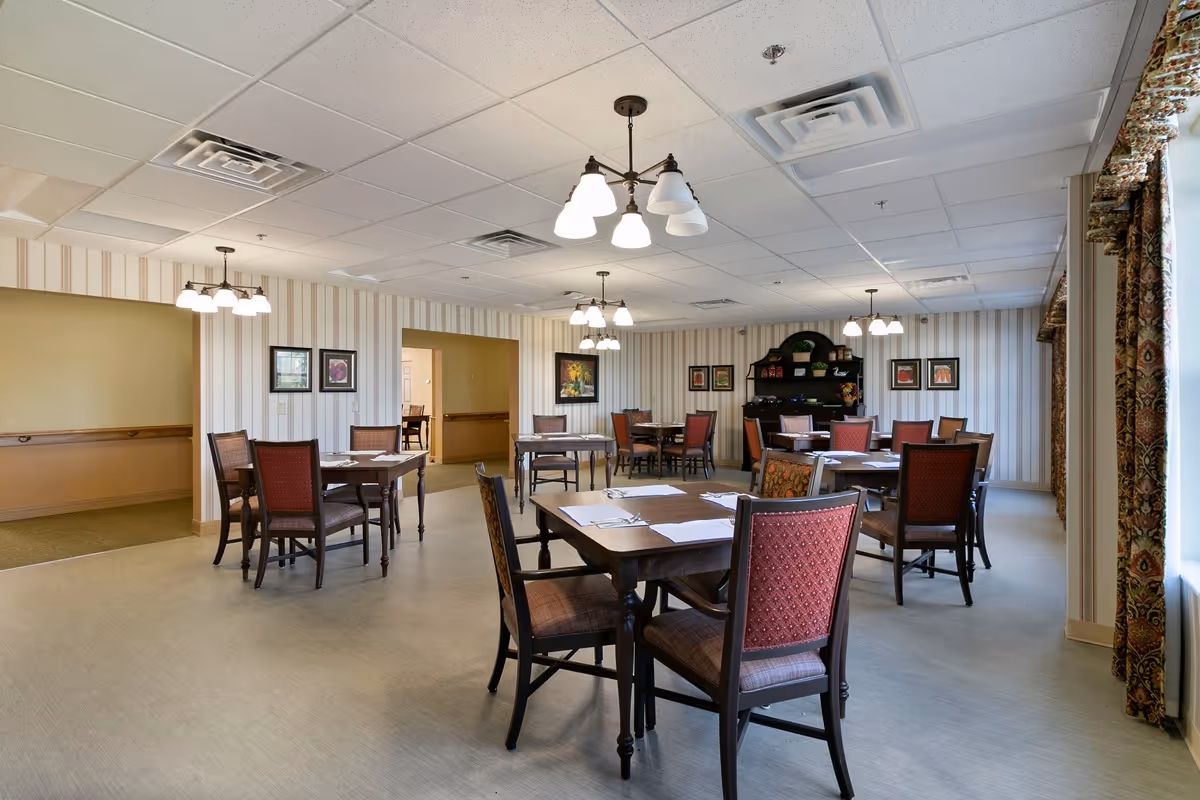 A dining room in St. Andrew’s Village with several wooden tables and chairs arranged neatly. Each table is set with placemats and silverware. The room has striped wallpaper, framed artwork on the walls, and multiple ceiling light fixtures. A window with patterned curtains allows natural light to enter the space.