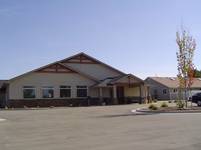 Exterior view of a single-story senior living facility building with beige siding, stone accents, and a covered entrance. There is a paved driveway and a small landscaped area with a young tree and shrubs under a clear blue sky.