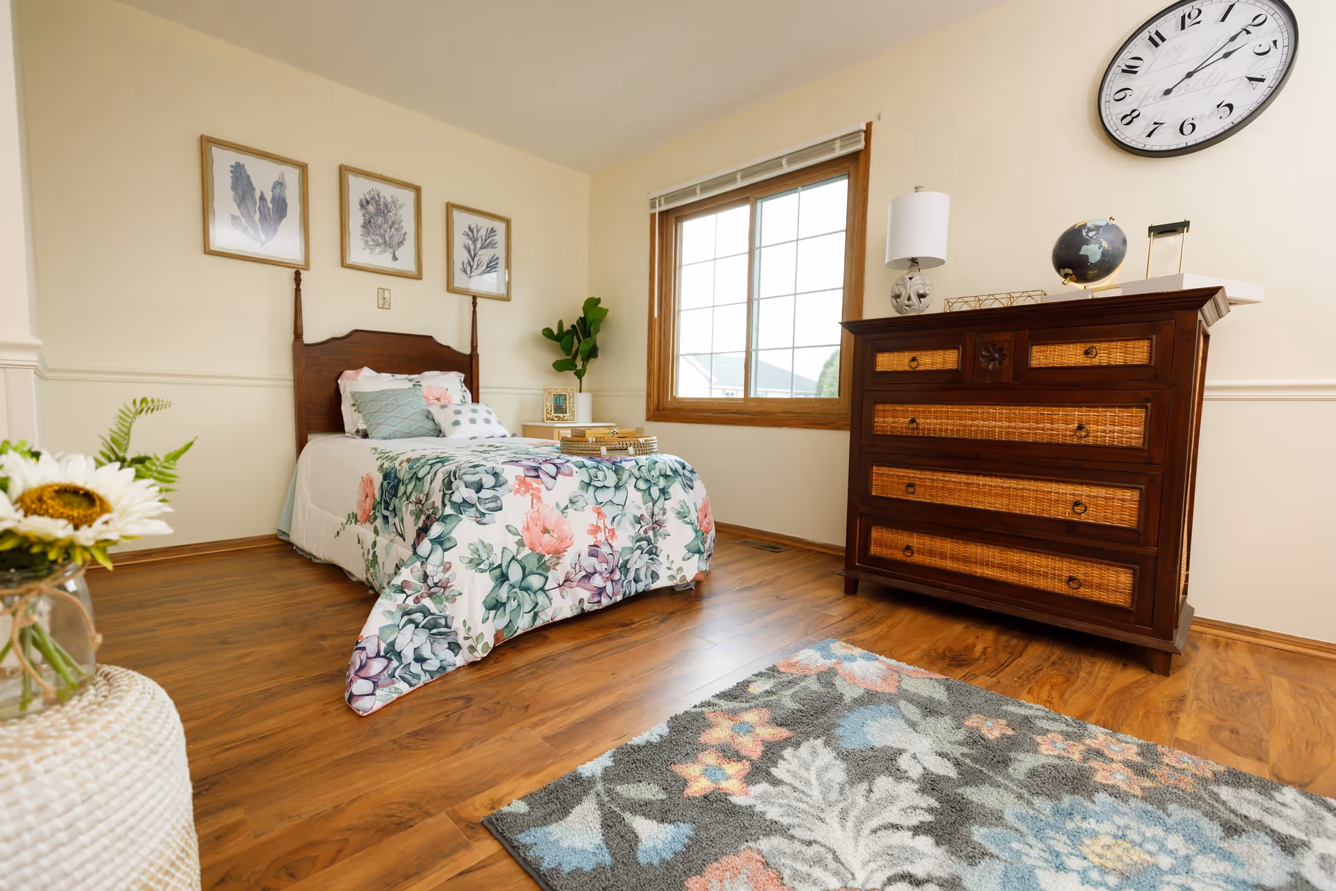 A bright bedroom with a single bed dressed in floral bedding, a wooden dresser, a large window, and hardwood floors.