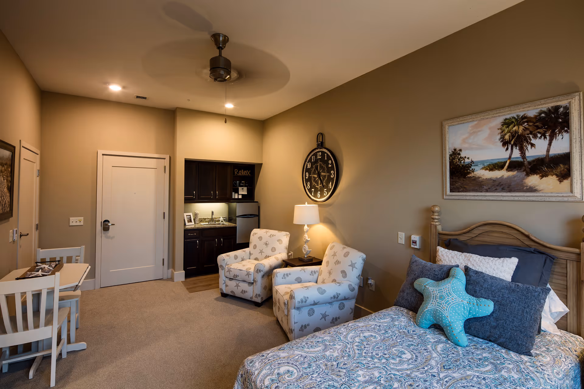 A cozy bedroom in a senior living facility featuring a bed with patterned bedding and a starfish-shaped pillow, two upholstered armchairs with a sea-themed pattern, a small kitchenette with dark cabinets and a mini fridge, a wall clock, a table with chairs, and a ceiling fan. The walls are painted beige and decorated with beach-themed artwork.