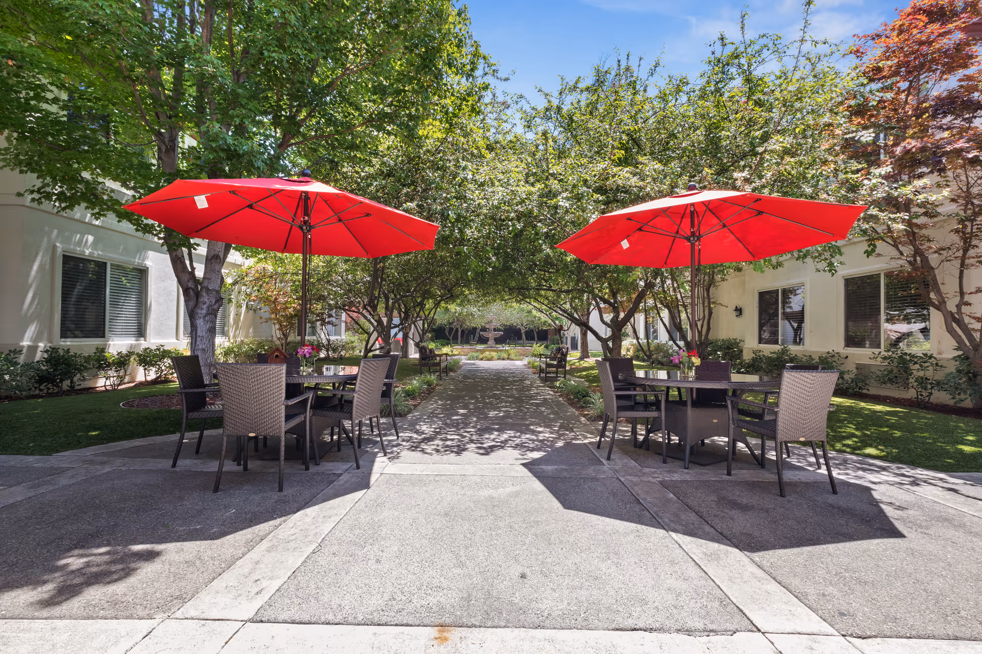 Courtyard with outdoor dining tables and bright red umbrellas along a tree-lined walkway between buildings.