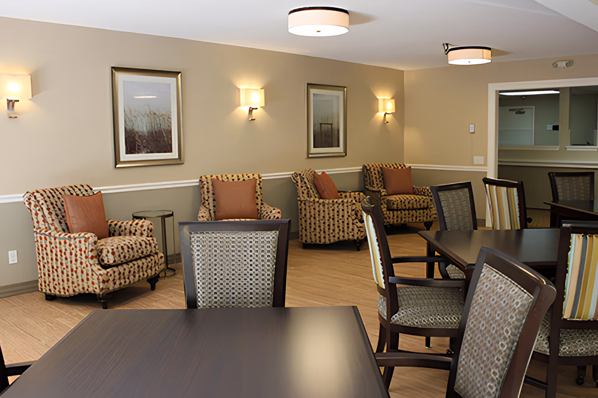 A furnished senior living common area with dining tables and upholstered armchairs against beige walls.