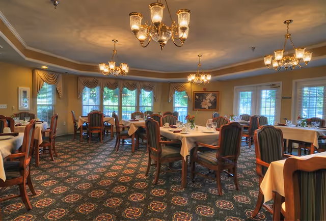 A spacious dining room with multiple tables covered in white tablecloths, each set with glassware and small floral centerpieces. The room features patterned carpet, large windows with draped curtains, and elegant chandeliers hanging from the ceiling providing warm lighting.