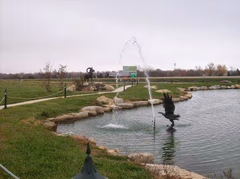 A landscaped outdoor area featuring a pond with a water fountain and a sculpture of a bird in flight. The pond is bordered by rocks and grass, with a walking path and additional sculptures visible in the background under an overcast sky.