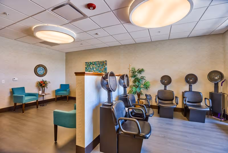 Interior salon area with multiple hooded hair dryers, black styling chairs, and a seating nook with teal armchairs under large ceiling lights.