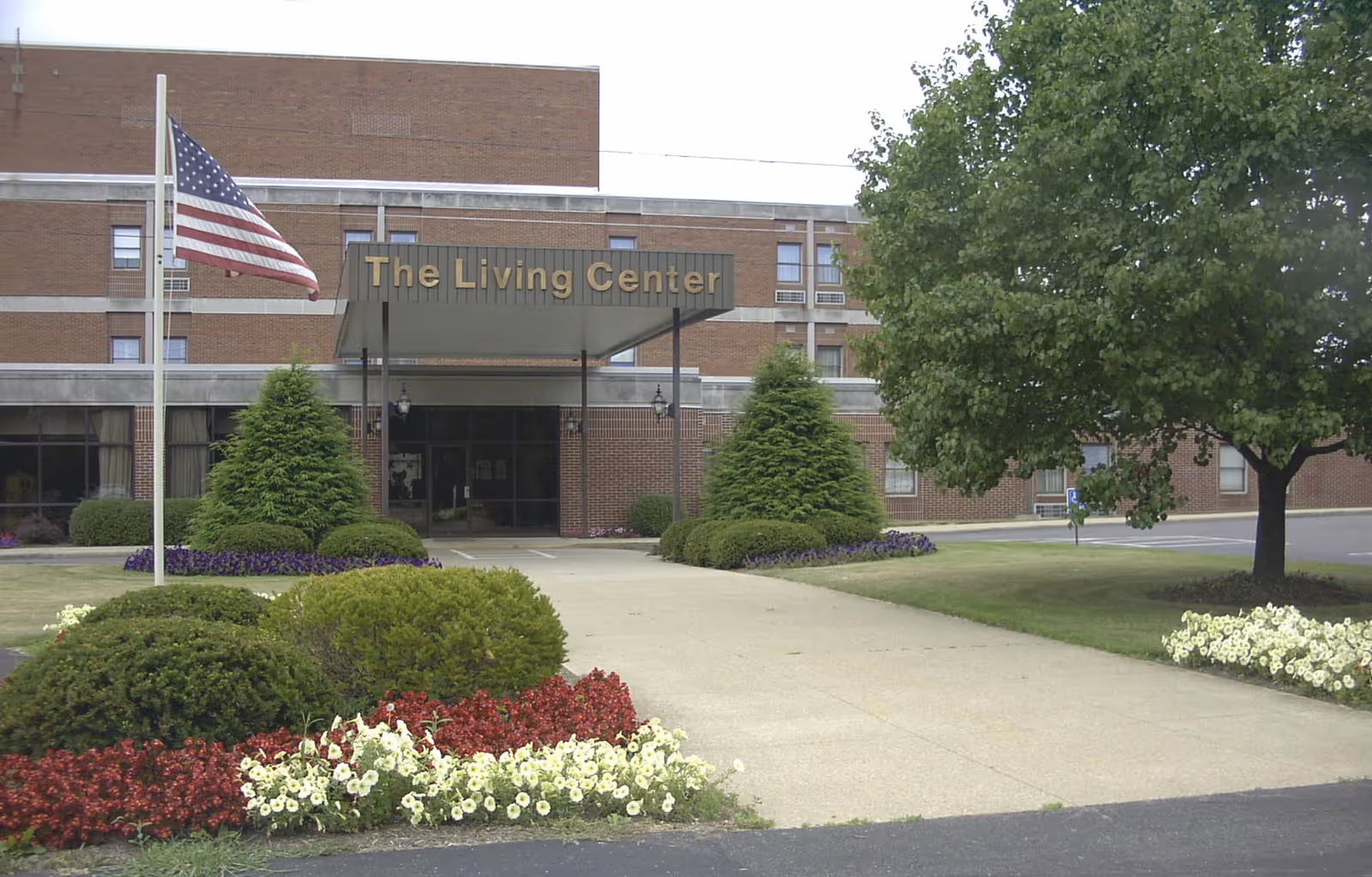 Front entrance of a brick building with a covered entry sign reading "The Living Center", landscaped shrubs, flowers, and an American flag.