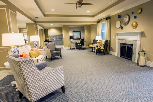 A spacious and well-lit living room area in an assisted living facility featuring patterned armchairs, a sofa with colorful cushions, table lamps, a fireplace with decorative plates above it, a desk with a chair, and a ceiling fan.