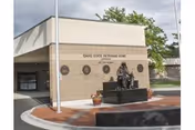 Exterior view of the Idaho State Veterans Home - Lewiston building entrance with a statue of a seated soldier in front, surrounded by a brick walkway and some greenery in the background.