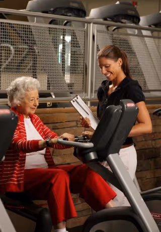 An elderly woman in a red and white outfit is using a seated exercise bike in a gym or fitness area, while a younger woman in a black shirt and white pants stands beside her holding a clipboard and smiling.