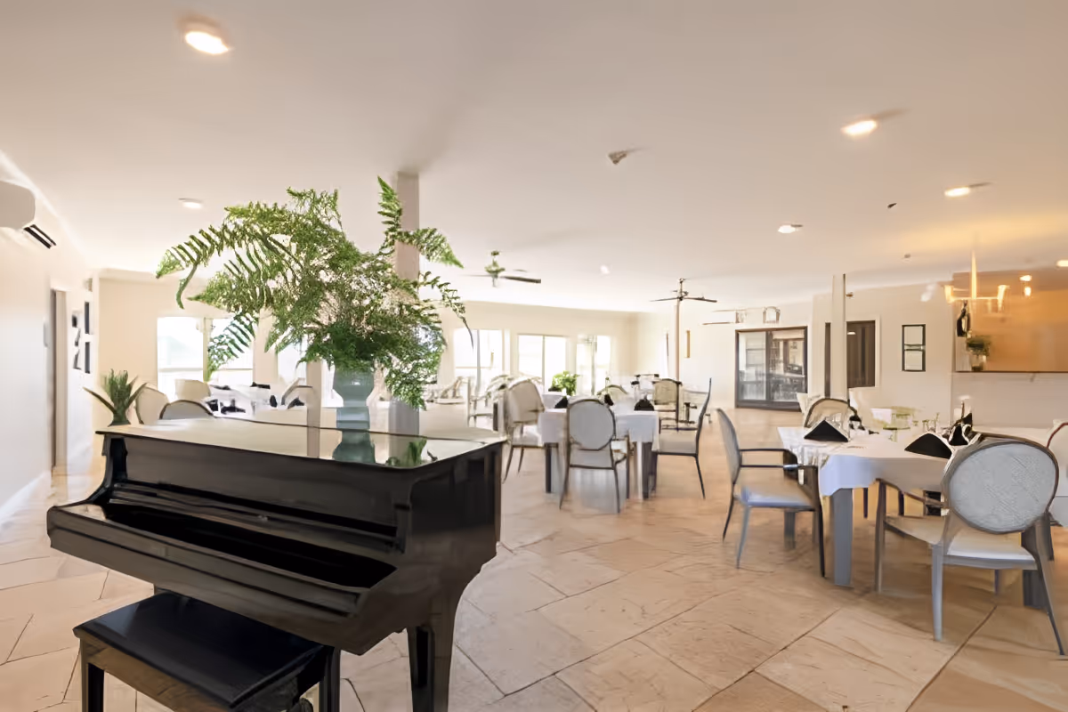 Bright and spacious dining room with multiple tables covered in white tablecloths and set with black napkins. A black grand piano with a large green fern plant in a vase on top is in the foreground. The room has beige tiled floors, white walls, ceiling fans, and large windows letting in natural light.