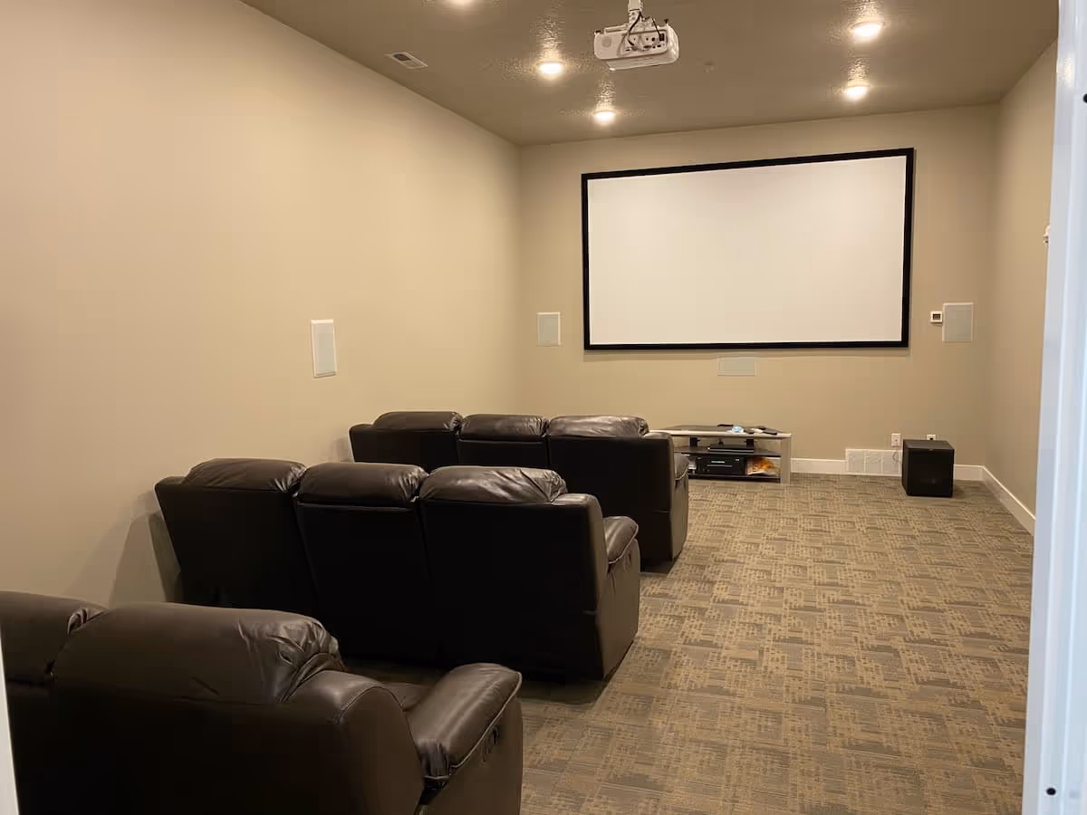A small home theater room with beige walls and carpeted floor. There are two rows of black leather recliner chairs facing a large white projection screen mounted on the wall. A projector is mounted on the ceiling, and a small media console with electronic equipment is placed below the screen.