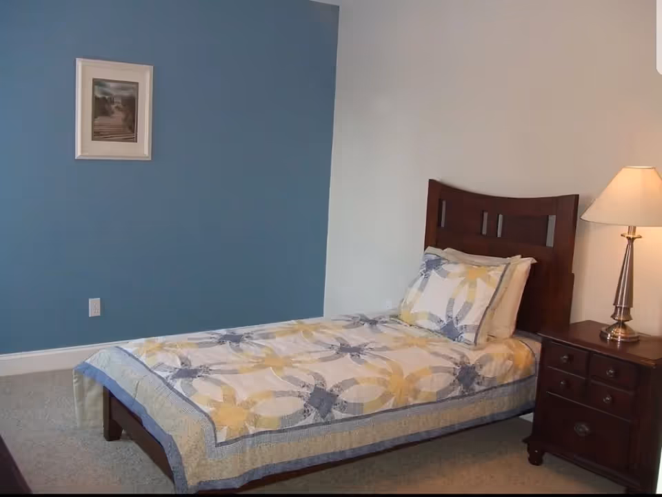 A bedroom with a single bed featuring a patterned quilt and matching pillow. The bed has a dark wooden headboard. Next to the bed is a dark wooden nightstand with drawers, topped with a silver lamp with a white lampshade. The walls are painted blue and white, and there is a framed picture hanging on the blue wall.