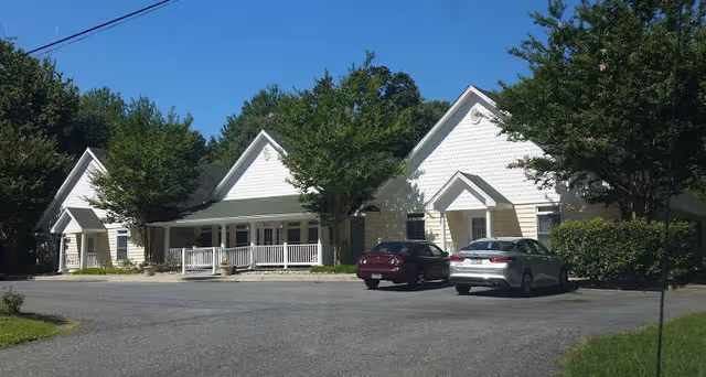 Front view of a single-story assisted living building with a white porch, peaked gables, trees, and two cars in the parking lot.