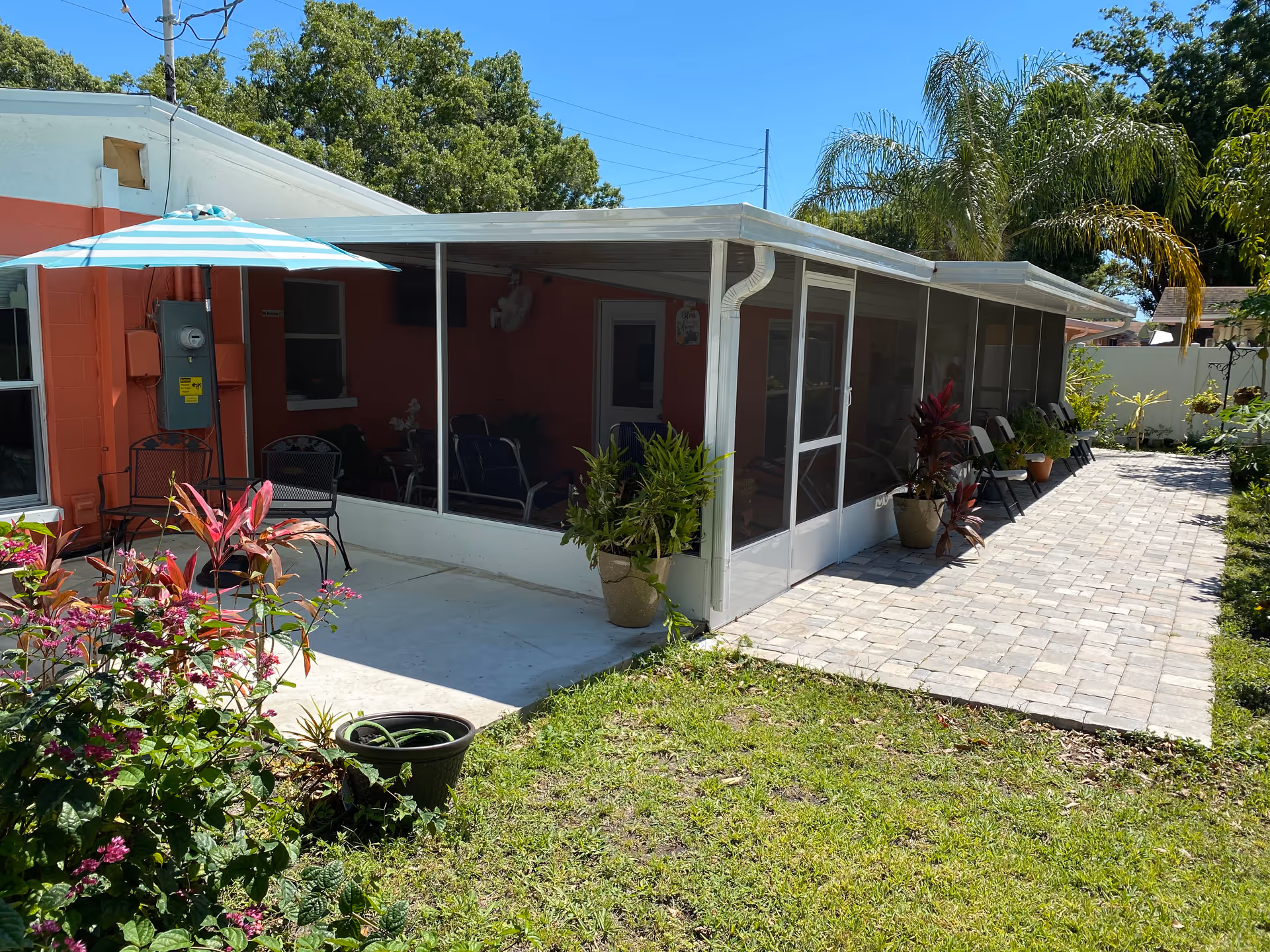 Screened porch and patio with potted plants, chairs and an umbrella beside a coral-colored building under a clear blue sky.