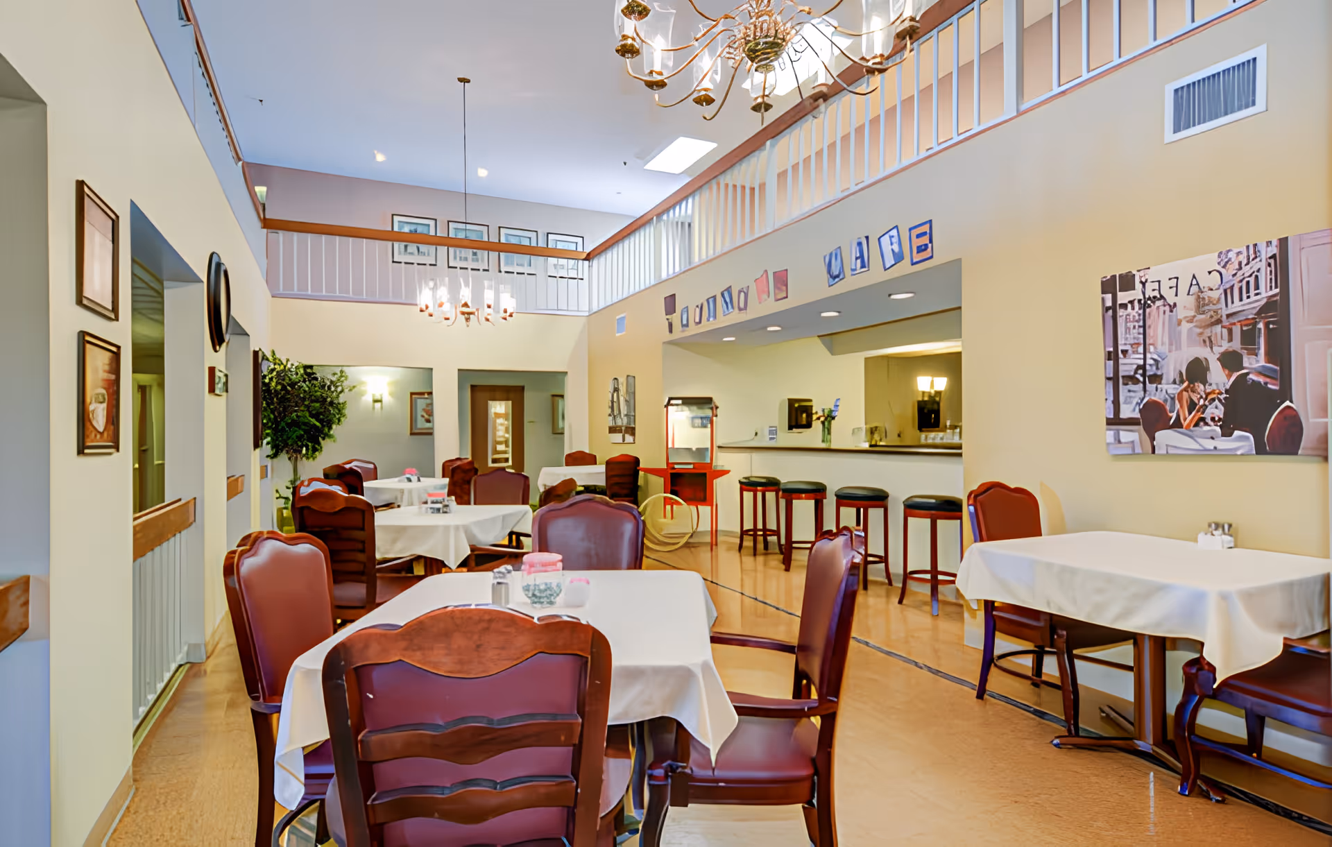 Bright senior living dining area with tables covered in white tablecloths, wooden chairs, a serving counter with stools, and a mezzanine level.