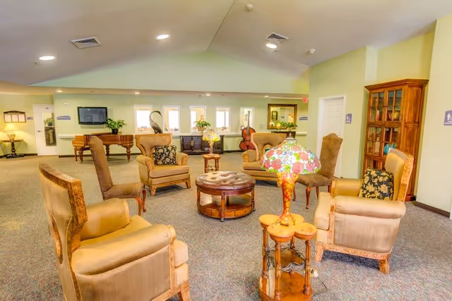 A spacious and well-lit common area with multiple upholstered armchairs arranged around a central round ottoman. The room features a vintage-style stained glass lamp on a small wooden side table, a wooden cabinet with glass doors, a piano, and a cello near the back wall. The walls are painted light green, and there are several windows letting in natural light.
