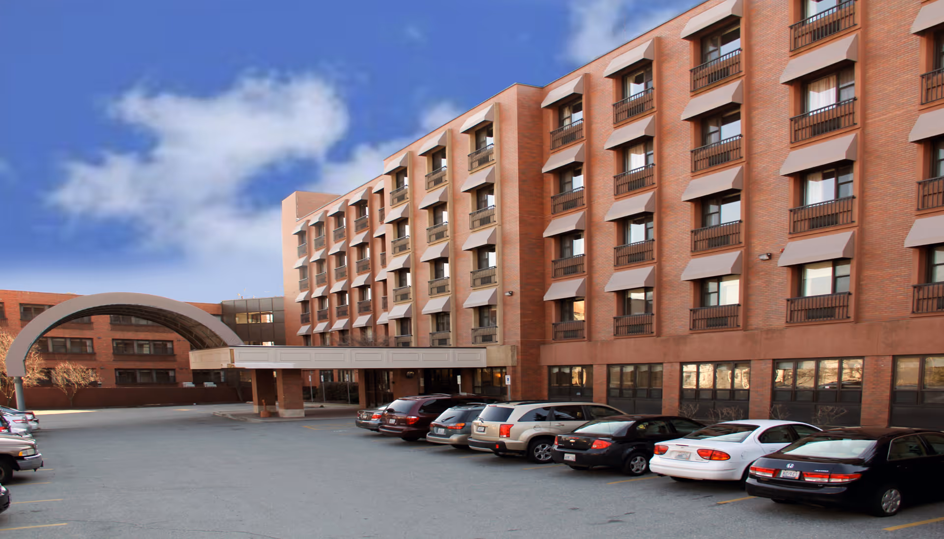 Exterior view of a multi-story brick building with several rows of windows, a covered entrance, and a parking lot with multiple parked cars under a partly cloudy blue sky.