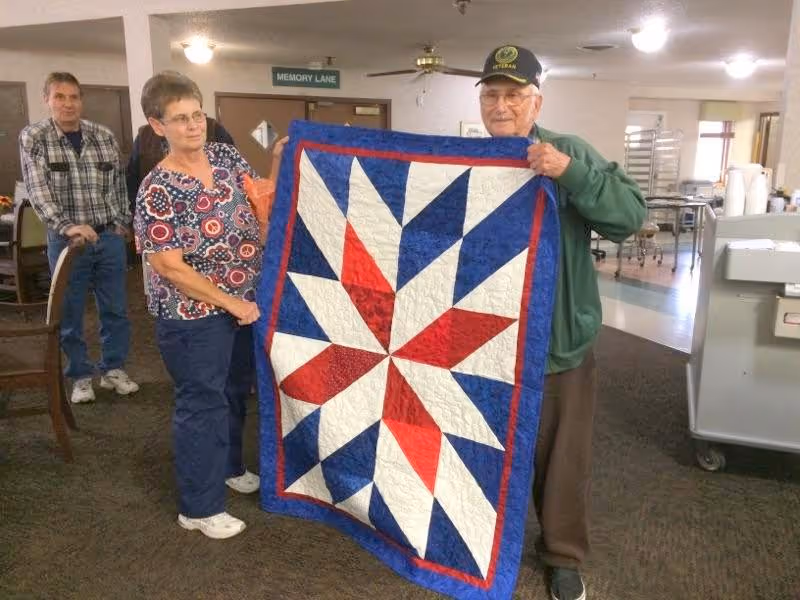 Two elderly individuals holding up a colorful quilt with a star pattern in red, white, and blue inside a senior living facility. In the background, a man stands near a chair, and there is a sign on the wall that reads 'MEMORY LANE'. The room has carpeted flooring and various furniture and equipment.