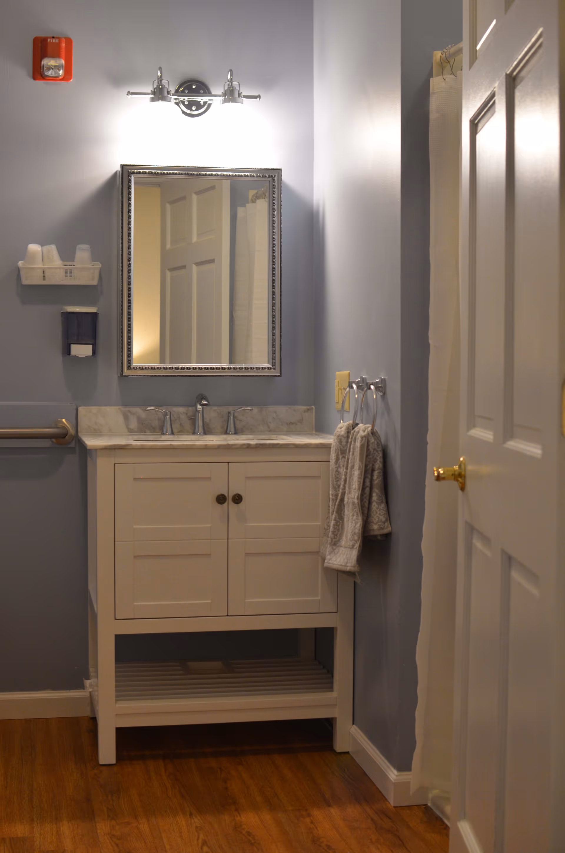 A bathroom vanity with a white cabinet and marble countertop, a rectangular mirror with a decorative frame above it, and a light fixture with two bulbs mounted on the wall. There are two hand towels hanging on a towel ring to the right, a soap dispenser and a small shelf with cups on the left wall, a fire alarm above the shelf, and a partially open door on the right side. The floor is wooden and the walls are painted light gray.