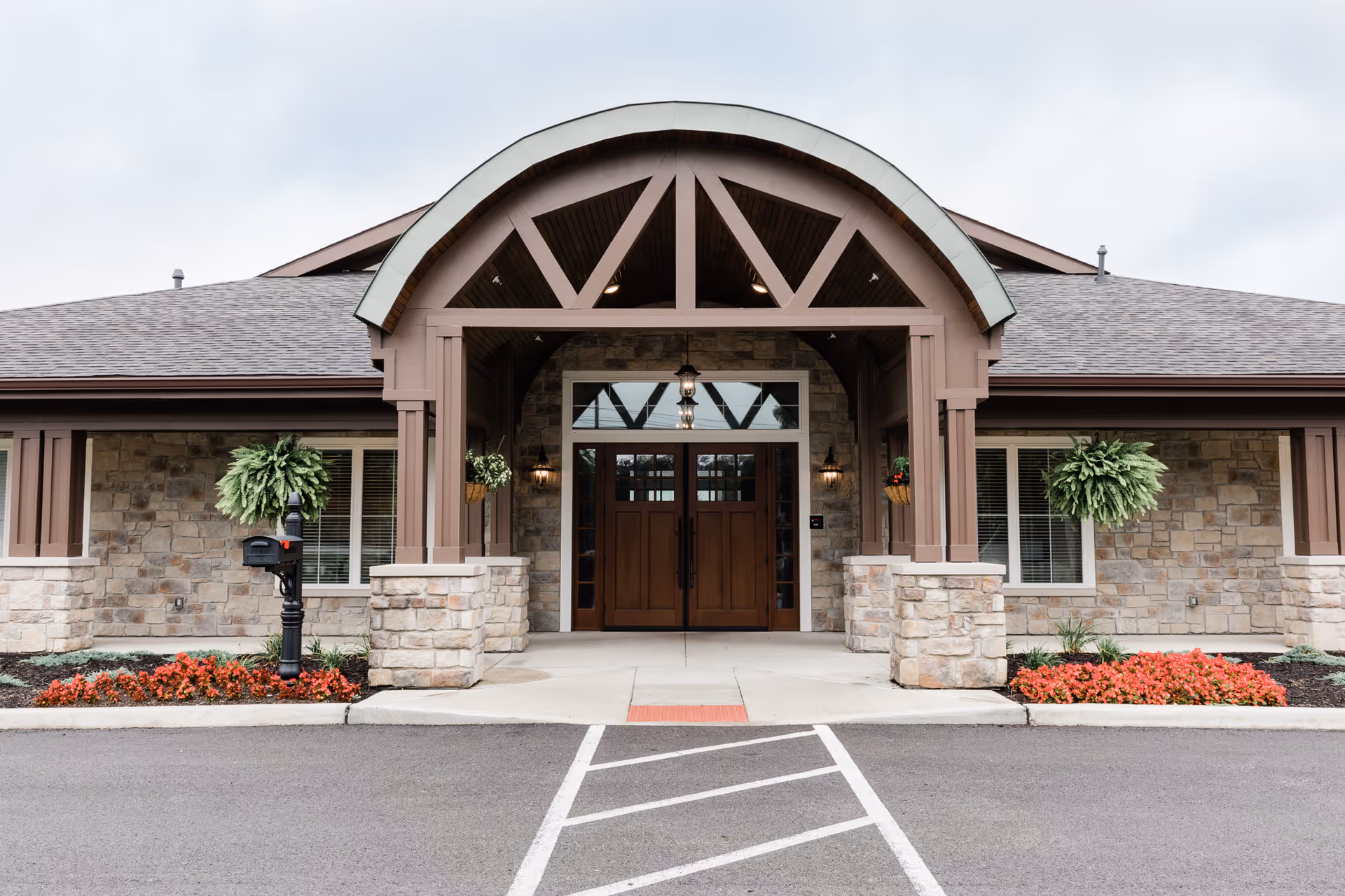 Front exterior entrance of a building with stone walls and a covered porch supported by wooden beams. There are two large wooden doors with glass panels, hanging plants on either side, and flower beds with red flowers along the front. A black mailbox is visible on the left side near the entrance.
